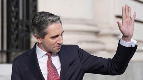 Simon Harris wearing a dark suit, with a white shirt and a red tie, waving. He is standing in front of a government building. 