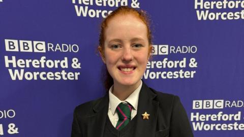 A teenage girl with red hair and wearing a white shirt, a green and red tie, a black v-neck top and a black school blazer, stands in front of a purple backdrop with the words "BBC Radio Hereford & Worcester" printed on it.