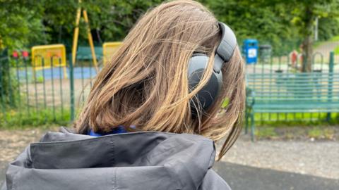 An anonymous child with short brown hair, outdoors, near a playground in a park, wearing headphones