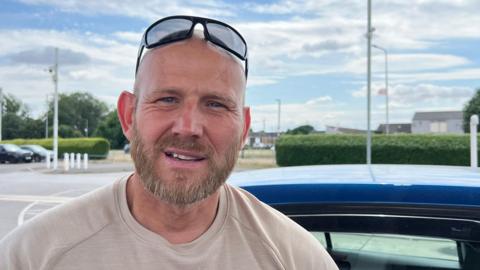 Wayne Parsons is a muscled and bald middle aged man with blue eyes and sunglasses on his head. He's wearing an immaculately laundered beige T-shirt and is standing beside his ultramarine car at a petrol station.