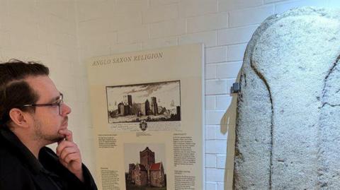 A man with glasses looks at a piece of art. Next to it is a board that says 'anglo saxon religion' 