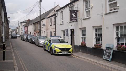 West Street. It is a quiet narrow lane lined with houses and on one side a row of cars are parked, with a police car parked at the front of it. There is a pub called The Thirsty Scholar that has window boxes of flowers and a chalkboard advertising a Sunday roast.