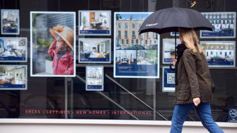 Woman walking past an estate agent window holding an umbrella.