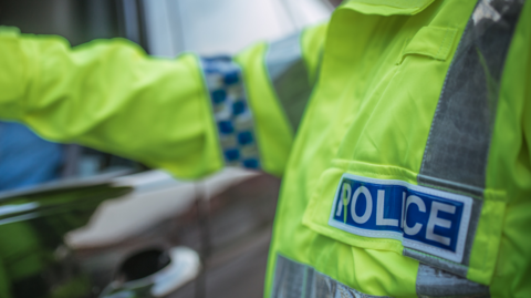 A police officer making a traffic stop in the street. The police officer is wearing a hi-vis jacket with "police" on it and putting his hand into the car.