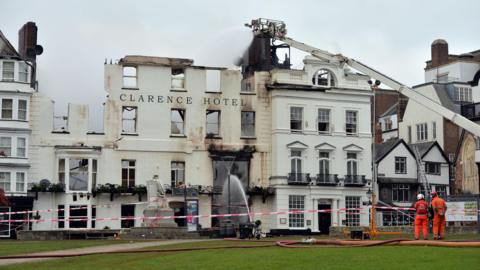 Firefighters in orange suits oversee water being sprayed from a crane onto the fire-damaged Royal Clarence Hotel. The four-storey historic building is partially burnt, with red and white tape cordoning off the area and a green lawn in the foreground.