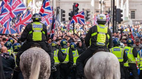 Police officers in helmets and high-visibility jackets, some on horseback, form a line facing a large crowd waving Union Jack flags during a rally.