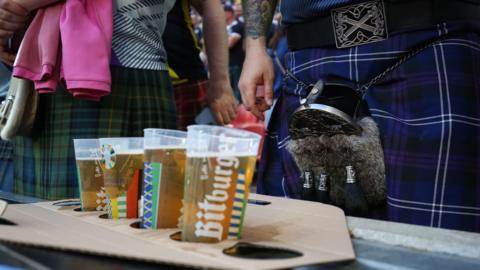 A cardhold drinks holder with four pints in it is in the foreground. In the background a man in a kilt can be seen.