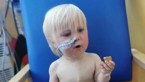 A young boy, with a tube in his nose and a sticker covering the tube. He has short blonde hair and blue eyes. He is holding a biscuit. The back of a blue hospital chair is behind him. 