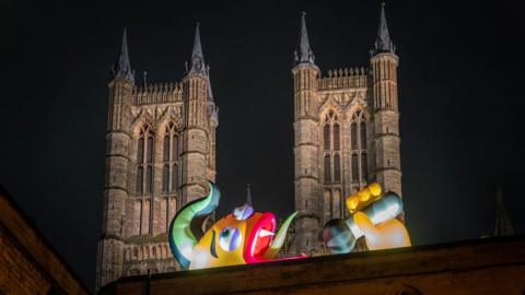 An illuminated yellow, green, purple and red inflatable monster on top of a wall and in front of two towers of a cathedral. It has a green tuft of hair and is lying on its side with its mouth open with a green snaky tongue and is holding a microphone in its yellow hand, mimicking being in mid-song.