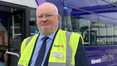 Piers Marlow wearing a high visibility First Bus vest and standing in front of a purple double decker bus 