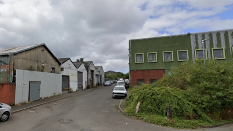 A row of units stand on the left side with a green building on the right. There are some cars parked in the road.