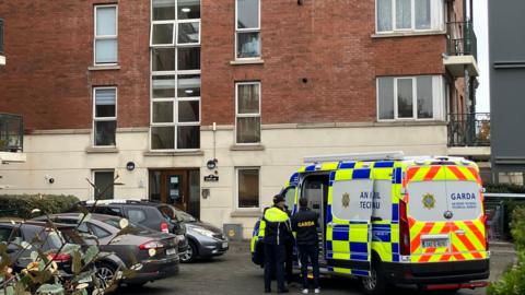 An apartment complex with cream bricks on the bottom and red bricks on the top half. There are cars parked to the left and a Garda van to the right. There are two gardaí standing beside the van. 