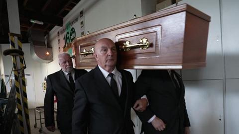 Four men carrying a wooden coffin with brass handles inside a funeral home. They all wear black suits.