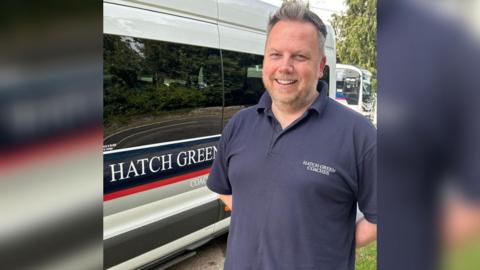 Nick is stood on the right-hand side of the picture, arms behind his back, smiling at the camera. He is stood next to one of his white minibuses, which says 'Hatch Green' on the side.