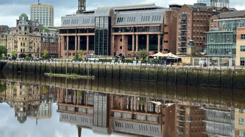 An imposing court building made of red stone and columns with large dark windows reflected in a river running in front of it