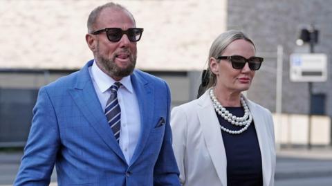Derek Hatton with short brown hair and beard a blue suit and light blue shirt and grey and navy stripy tie walks with his wife Sonjia Hatton who has blond hair  tied back wearing sunglasses and a white blazer, navy t-shirt and a pearl necklace.