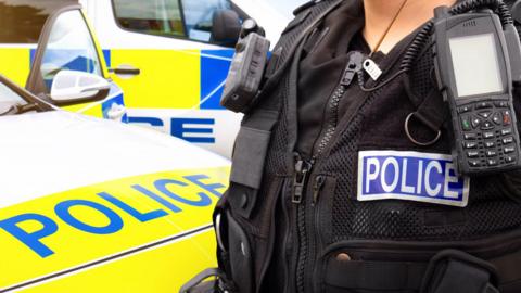 The front of a police car and an officer standing next to it. On the officer's chest is a blue strip saying "police" in white behind a walkie-talkie. There is another police car behind the first