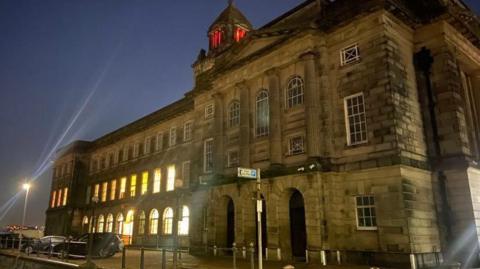 Wallasey Town Hall is seen at night, with the lights on in a number of windows on the left hand side. Some cars are parked at the side of the building.