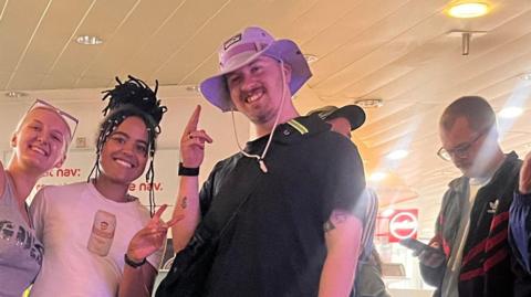 Laurence wears a black T-shirt, black shorts and a purple hat. He stands next to his friends Meg (left) and Kitty (middle) in the coach station surrounded by bags. They smile at the camera as Meg and Kitty do peace signs