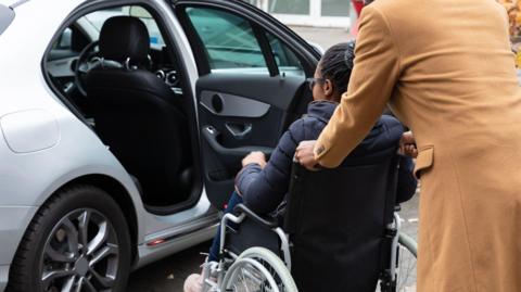 A parent helping their child who is in a wheelchair getting into a silver car.