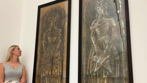 A woman in a grey vest top with long blonde hair stands beside two large artworks looking at them