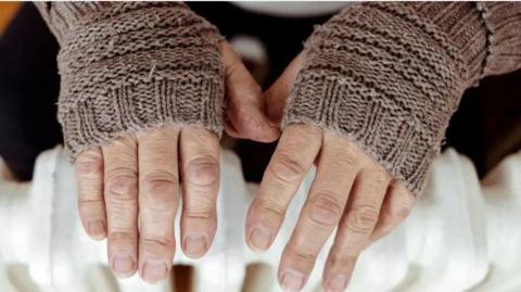 Close up of a pair of hands outstretched over a radiator wearing fingerless gloves 