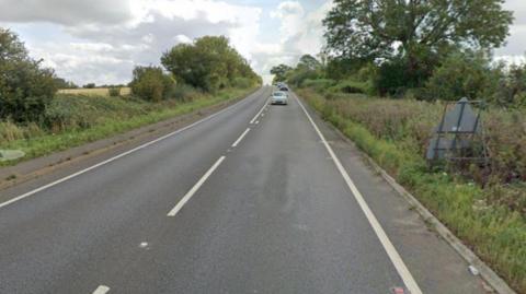 A street view of the A5 single carriageway - a long straight single carriageway with grass verges and hedges on either side. Several cars are driving up the road in the distance.