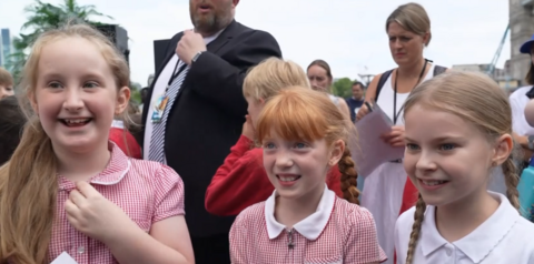 Three girls wearing red and white school uniforms. They are standing next to each other and looking to the side of the camera. They are all smiling.