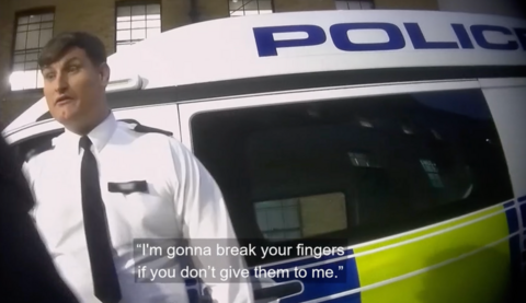 A screenshot from undercover filming showing an officer in a uniform made up of a white shirt and a black tie standing in front of the side of a police vehicle. A subtitle on the image reads "I'm gonna break your fingers if you don't give them to me."