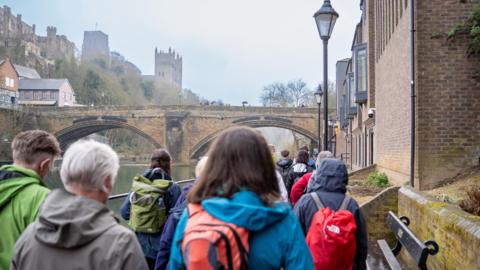 Back view of the head and shoulders of a group of walkers on a path with buildings to one side and a river on the other. They are wearing anoraks or kagoules and some have small backpacks. Ahead of them an arched stone bridge spans the river and Durham Cathedral and Castle stand to one side above it.