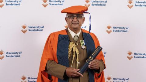 Ben Banerjee receiving his honorary doctorate of medicine from the University of Sunderland. He is wearing an orange academic cap and gown as well as a brown military uniform. He is holding a scroll and looking into the camera.