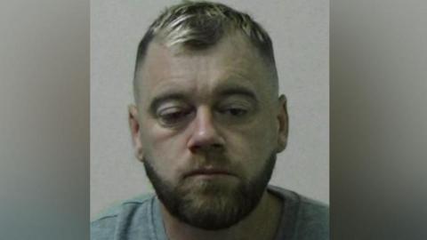 Mugshot of Steven Rycroft. He has short dark hair with blond highlights and a brown beard. His right eye is partially closed and he looks like he is dazed. He is wearing a grey shirt.