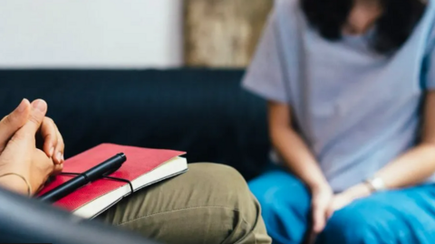 A person resting their hands on a notebook cross-legged, and a woman pictured in the background out of focus.