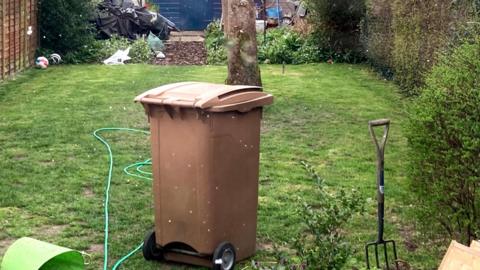 A brown wheelie bin, placed on the grass in the garden. 