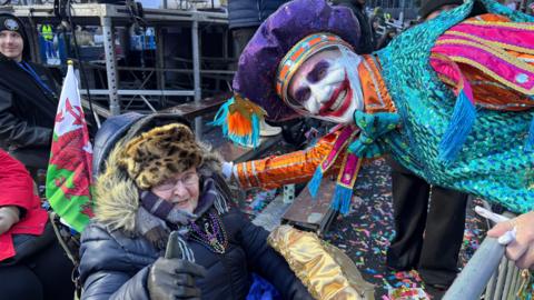 Photograph of Avril Davidge, sat outside in her wheelchair. A Welsh flag can be seen poking out of her wheelchair. She wears a leopard furry hat, a purple scarf and a black puffer coat. She wears small reading glasses and holds her thumb up to the camera. A captain of the Mummers parade leans down to her and smiles. He wears a vibrant blue, purple and orange sequined costume and has a large red joker smile painted on his face. 