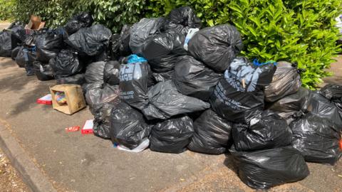 A large pile of black bin bags on a pavement. Some cardboard boxes have also been left.