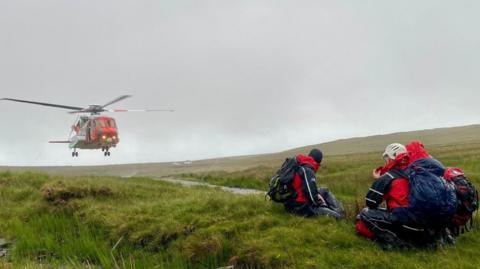 SLMRT responders awaiting the departure of Irish Coast Guard helicopter, Rescue 118. There are three team members in navy and red waterproofs, wearing hats and backpacks. They crouch on the grass. Mountains are visible in the background. The sky is cloudy.
