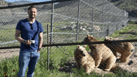 Steven Cree wearing jeans and a t-shirt, posing for the camera beside a wire fence. Three light brown bears are sat on the other side of the fence in a large enclosure.