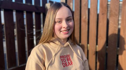 A smiling Niamdh looks into the camera lens. She has long dark blonde hair and is wearing a beige hoodie with "Proud to be deaf" embroidered on the front. She stands in her back garden with a brown wooden fence in the background.