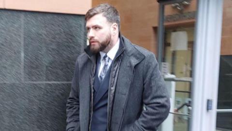 A man with short dark hair is pictured walking out of a building. He has a dark beard and wears a navy suit, white shirt and patterned tie and a grey overcoat.