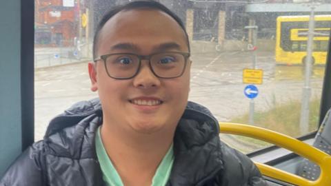 Maxson Goh smiles in a black coat and green shirt sat on the 651 bus. He is wearing glasses and can be seen at in front of the bus window on a rainy day. 