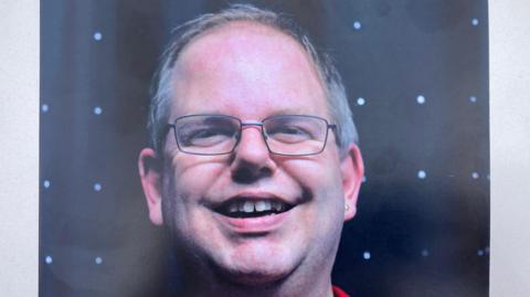 Jeremy Huggett wearing thin rimmed glasses smiling at the camera against a black background