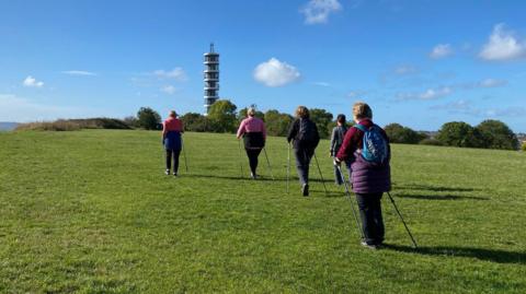 Five people facing away from the camera are walking up a hill at Stoke Park. They all have two poles. A satellite tower, Purdown BT Tower, is seen in the distance, near some trees.