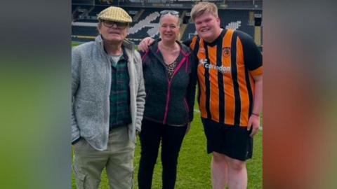 A boy on the right of three is wearing a Hull City kit and standing on a football pitch with his arm round his mum and dad. His dad has a grey zipped fleece with a flat cap and mum is wearing a dark grey and pink zipped hoodie.