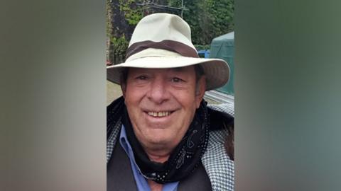 An older man smiling at the camera while wearing a black scarf with a white hat, with a brown stripe around the hat