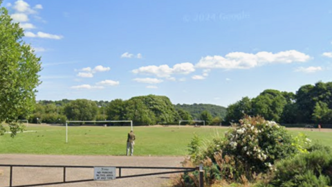 An area of grass with two sets of goalposts and surrounded by trees. In the foreground is a metal gate across an access road.