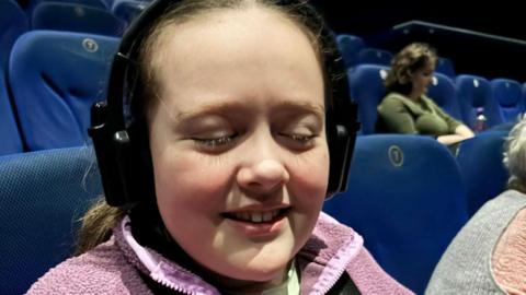 Eryn has long brown hair and is pictured at the cinema in a pink fleece and is wearing an audio descriptive headset. The cinema seats are all blue and a few people are seen in the cinema in the background. 