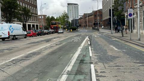 A city centre road with a bus lane on the right and several cars, a white van and a bus waiting at traffic lights on the left. Several brown and beige coloured buildings are either side of the road with a few people walking on the pavement.