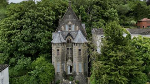 Wide drone image showing the large bricked synagogue surrounded by overgrown trees.