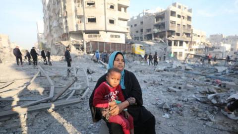 A woman sits with a child as Palestinians inspect buildings damaged in an Israeli air strike in Gaza City, northern Gaza (14 January 2025)